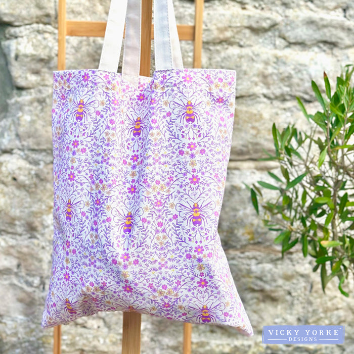 Tote bag with bee and floral pattern on a wooden stand against a stone wall.