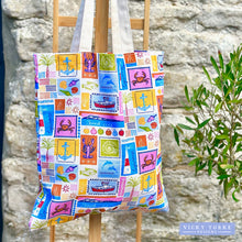 Load image into Gallery viewer, Close-up of an organic cotton tote bag covered in the &#39;Island Life&#39; pattern, a grid of colourful squares resembling vintage postage stamps, depicting coastal icons like a red boat, blue dolphin, lighthouse, lobster, and crab. The bag is hanging outside against a stone wall next to green foliage. The Vicky Yorke Designs logo is in the bottom left corner. 