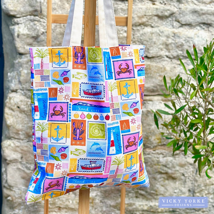 Close-up of an organic cotton tote bag covered in the 'Island Life' pattern, a grid of colourful squares resembling vintage postage stamps, depicting coastal icons like a red boat, blue dolphin, lighthouse, lobster, and crab. The bag is hanging outside against a stone wall next to green foliage. The Vicky Yorke Designs logo is in the bottom left corner. 